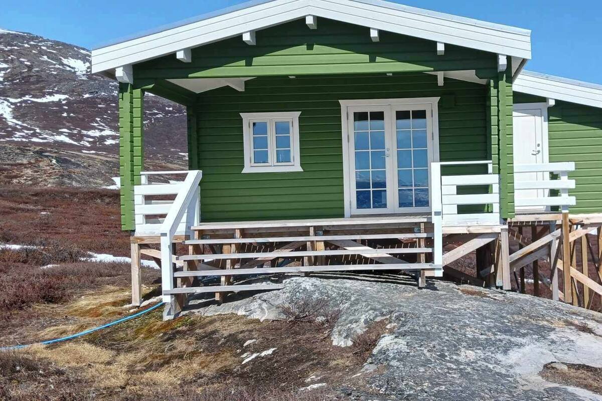 A green wooden cabin with a white porch and a single window, situated on a rocky outcrop near a body of water.