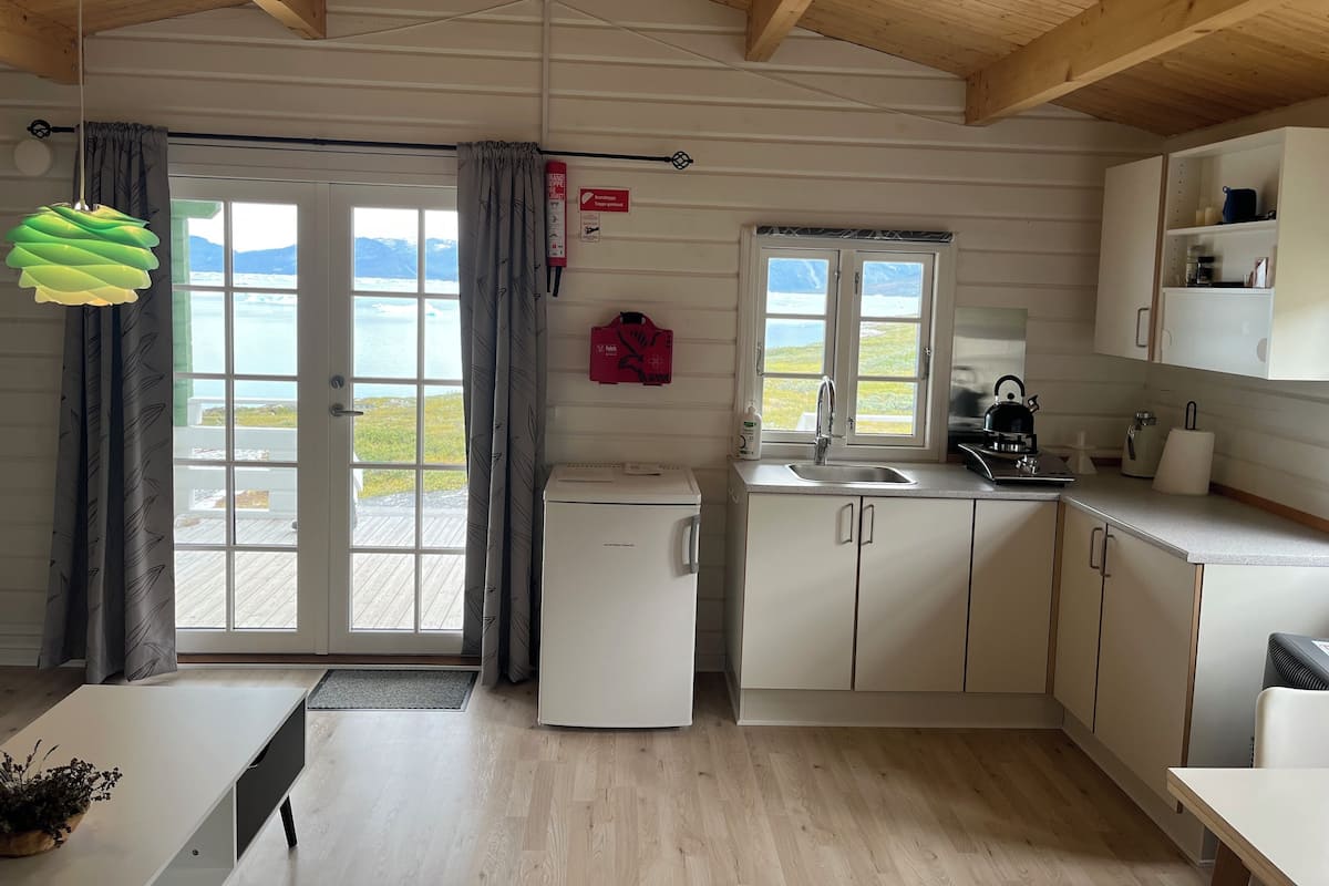 A compact kitchen with a refrigerator, sink, and cabinets, a window with a view of the outdoors, and a door leading to a balcony.
