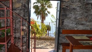 Ein raum mit steinernen Wänden, einer hölzernen Bank und einem Tisch, einer Wendeltreppe und Blick auf eine Palme sowie einen Strand durch eine offene Tür.