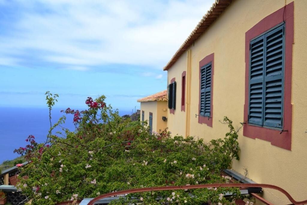 A coastal building with a balcony, a view of the sea, and a garden with flowering plants.