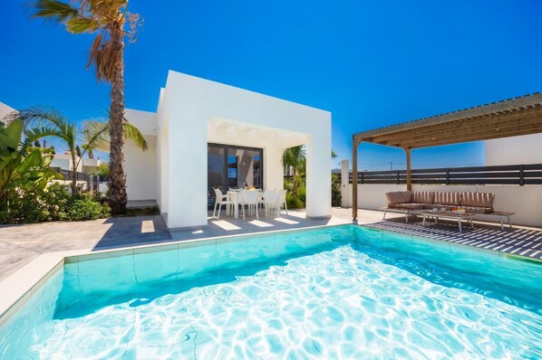 A modern outdoor pool area with a white structure, lounge seating, and palm trees.