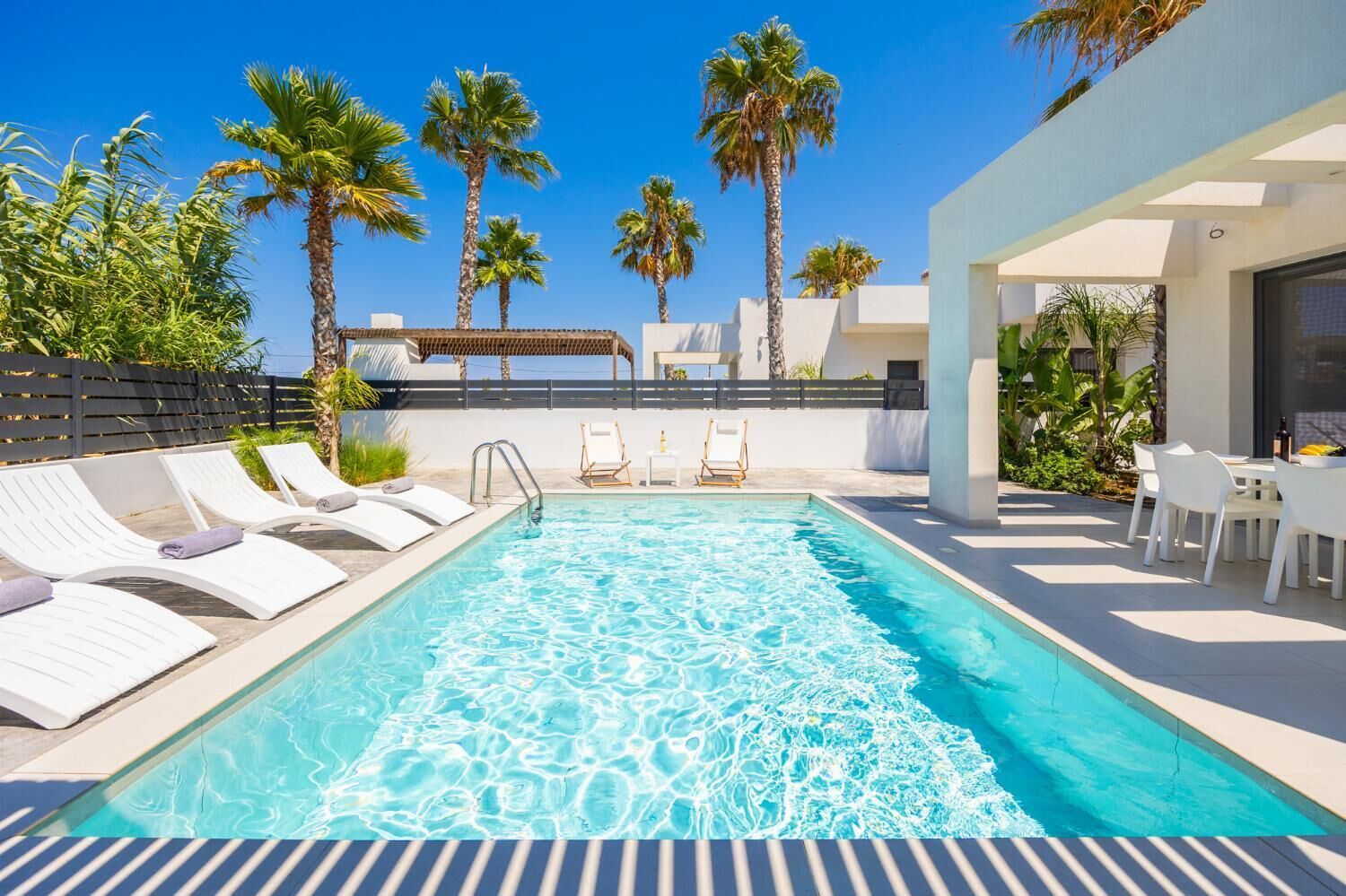 A swimming pool with lounge chairs and a table, surrounded by palm trees and a clear sky.