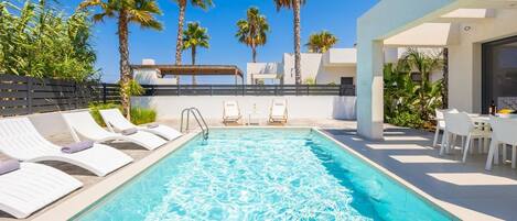 A swimming pool with lounge chairs and a table, surrounded by palm trees and a clear sky.