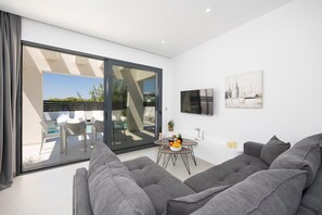 Un salon moderne avec un canapé gris, un téléviseur fixé au mur et un coin repas comprenant une table et des chaises.