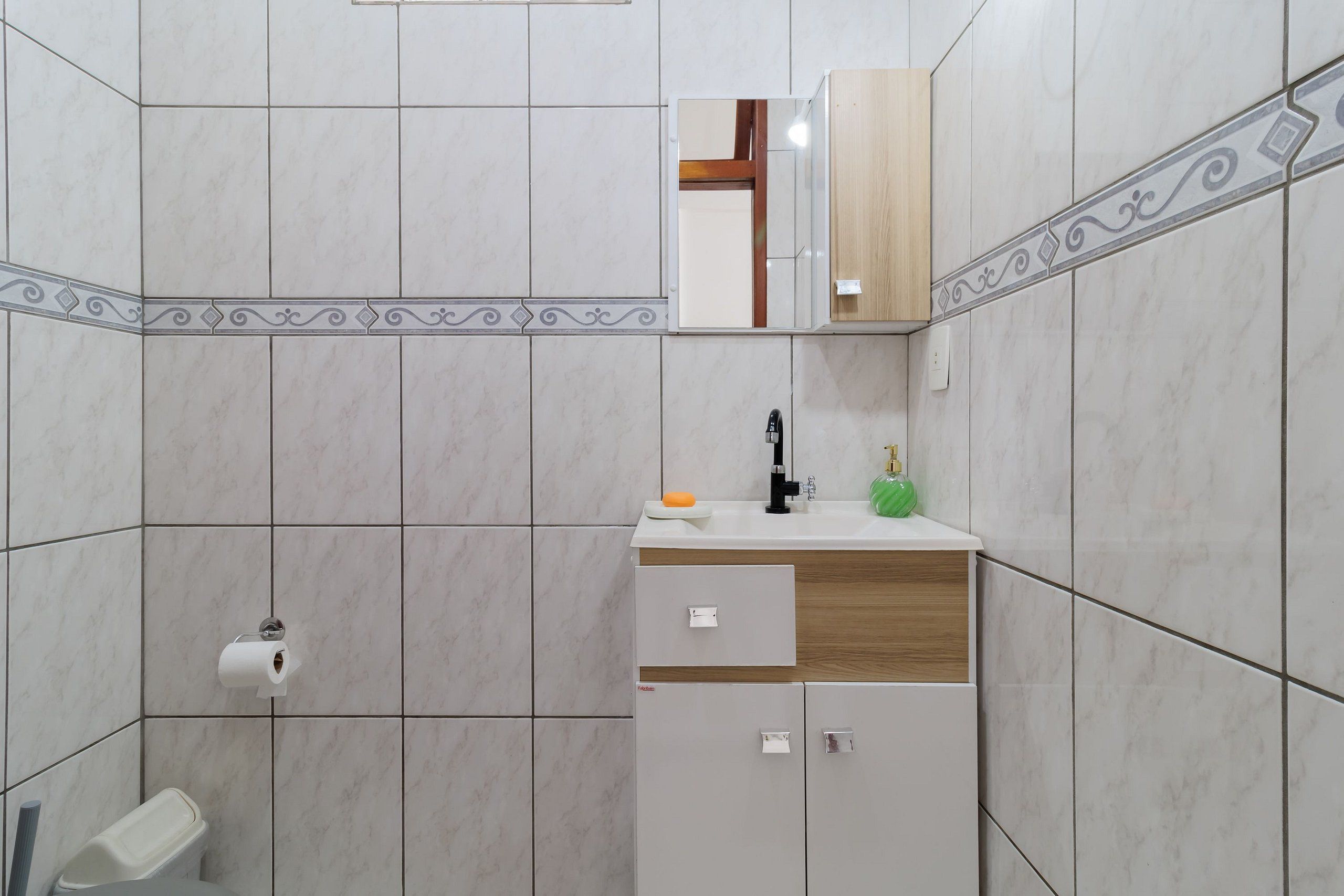 Un baño con baldosas de mármol blanco, un tocador de madera con un espejo, un inodoro y un lavamanos con jabón y una botella verde.