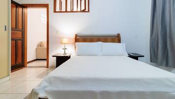 Una cama bien tendida con cabecera de madera, una mesita de noche con lámpara, y vista a una puerta que da a otra habitación.