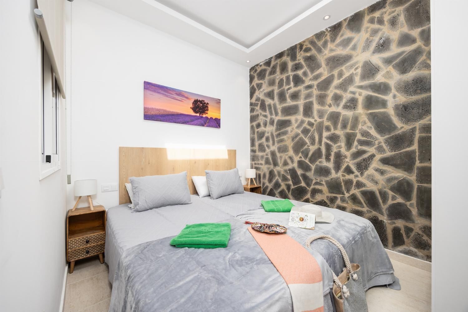 A bedroom with a stone wall, a bed with grey bedding, a green towel, and a painting of a sunset.