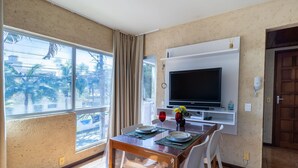 A dining area with a TV mounted on the wall, a table set with plates and glasses, and a window with curtains. - Apt 6 min From the sea in Canasvieiras-sc Rba201 (Florianópolis)