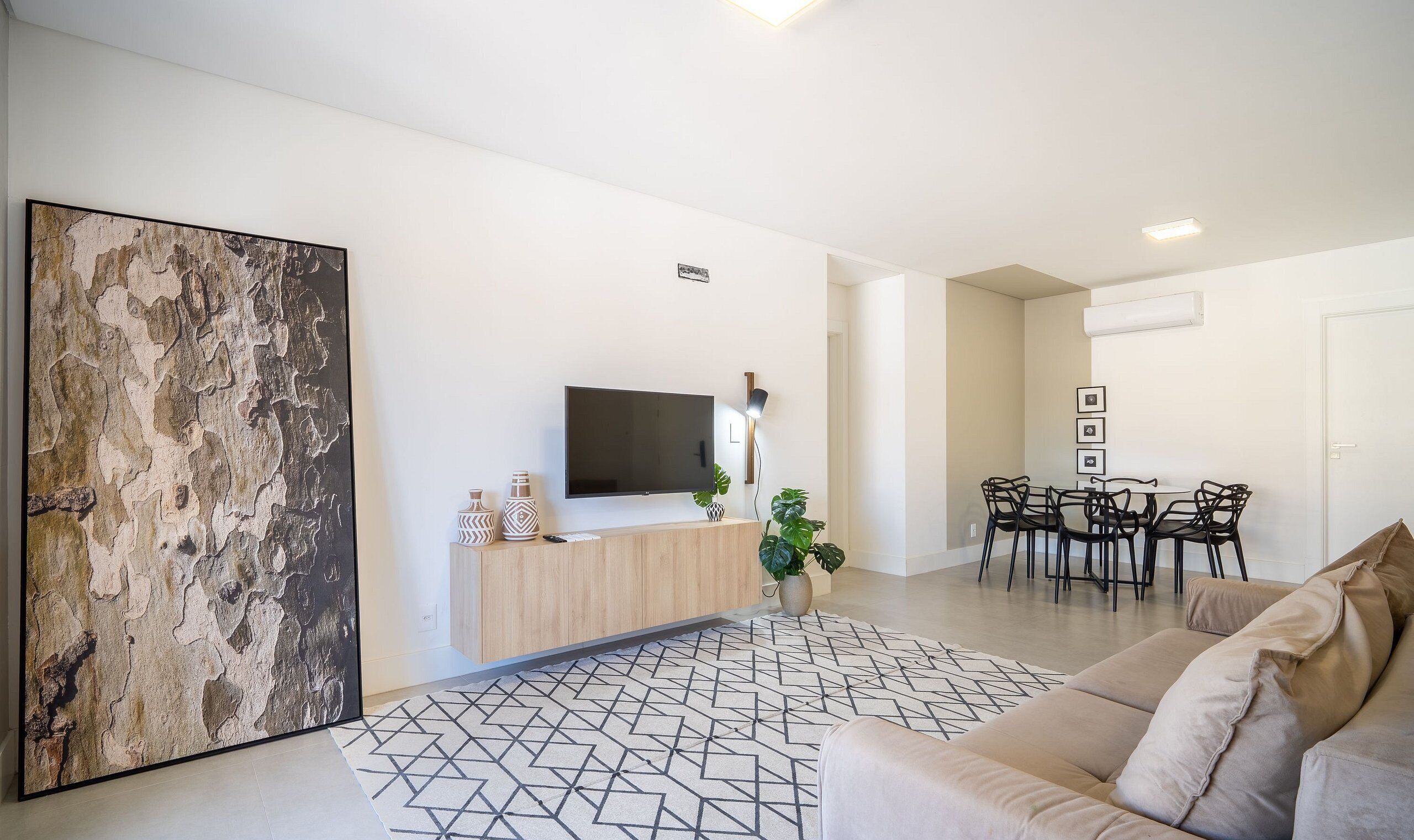 A modern living room with a large abstract painting, a flat-screen TV, a wooden console, a patterned rug, a beige sofa, and a dining area with a table and chairs.