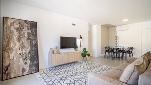 A modern living room with a large abstract painting, a flat-screen TV, a wooden console, a patterned rug, a beige sofa, and a dining area with a table and chairs. - Apartment w/ Barbecue 150m the Beach Jurere Ran203 (Florianópolis)