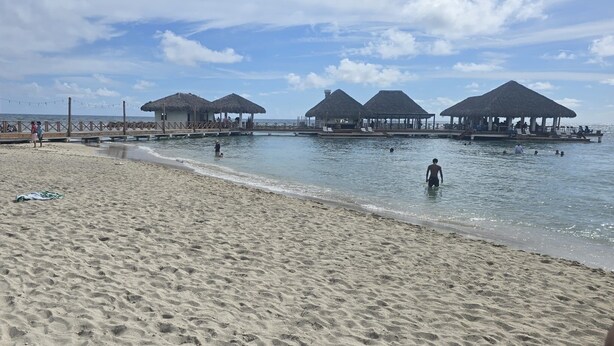 Une plage bordée de constructions aux toits de chaume, un rivage sablonneux et une mer calme.