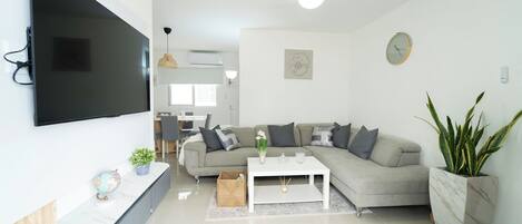 A modern living room with a flat-screen TV mounted on the wall, a grey sectional sofa, a white coffee table, and a large potted plant.