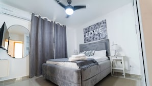 Une chambre à coucher moderne avec un lit gris, un ventilateur de plafond, un climatiseur fixé au mur, une table de chevet avec une lampe et un grand miroir.
