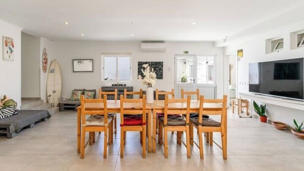 A spacious living area with a dining table, chairs, a sofa, a TV, and a surfboard. - Luxury private room with attached bathroom near the beach (Setúbal)