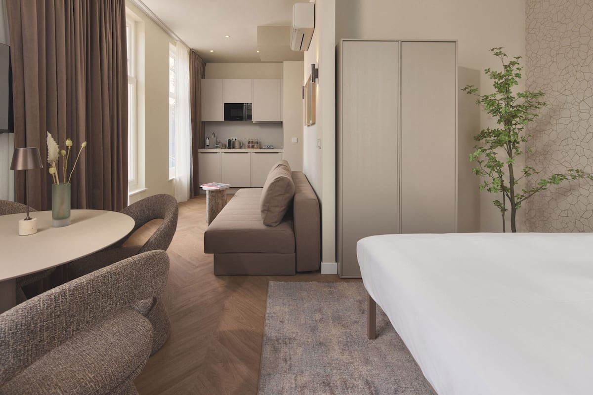 A modern hotel room with a sofa, a dining table, a bed, and a kitchen area in the background.