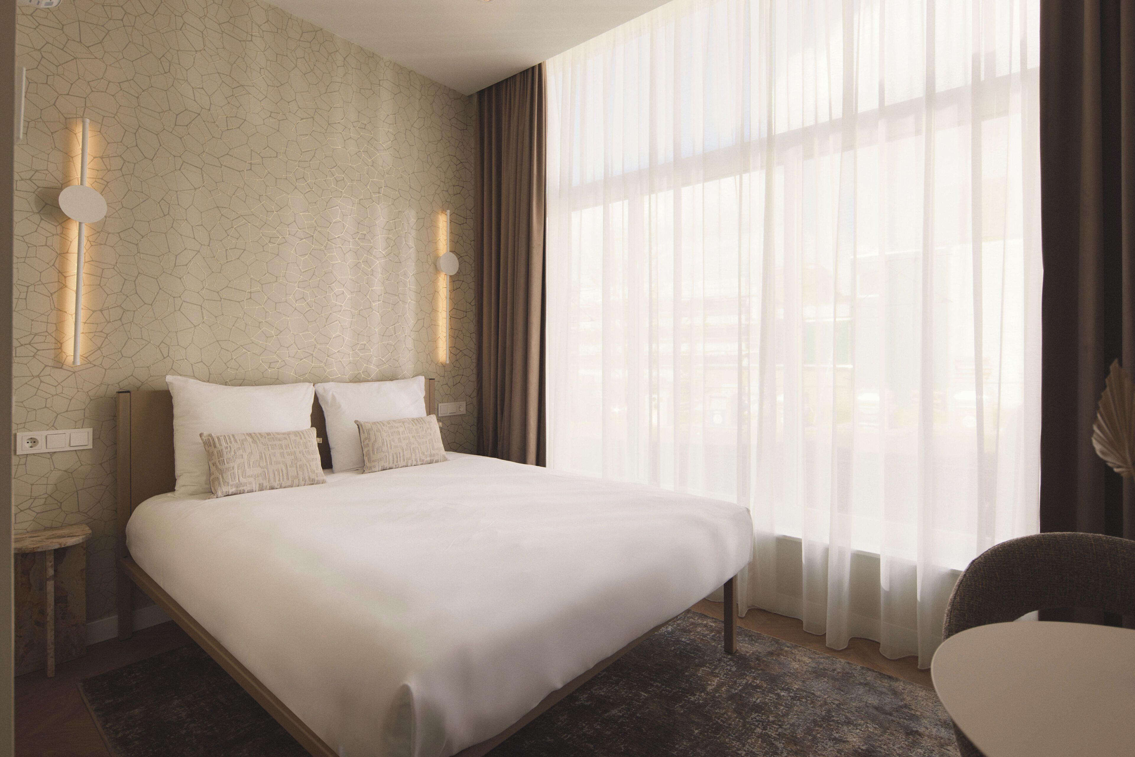 Une chambre d’hôtel avec un grand lit, une table de chevet, une chaise et une fenêtre avec des rideaux légers.