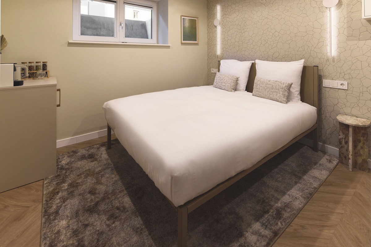 A bedroom with a bed, a small bedside table, a nightstand, a window, and a wall-mounted light fixture.
