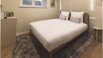 A bedroom with a bed, a small bedside table, a nightstand, a window, and a wall-mounted light fixture.
