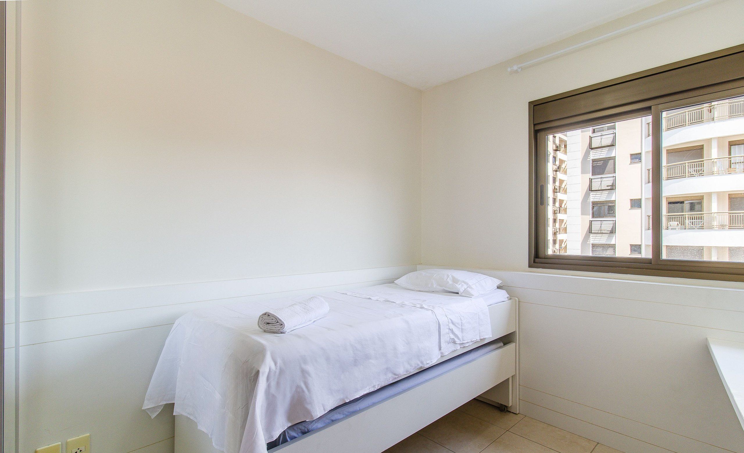 Una habitación pequeña y limpia con una cama individual, una ventana que da a un edificio y una pared blanca.