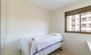 Een kleine, schone kamer met een enkel bed, een raam met uitzicht op een gebouw en een witte muur.