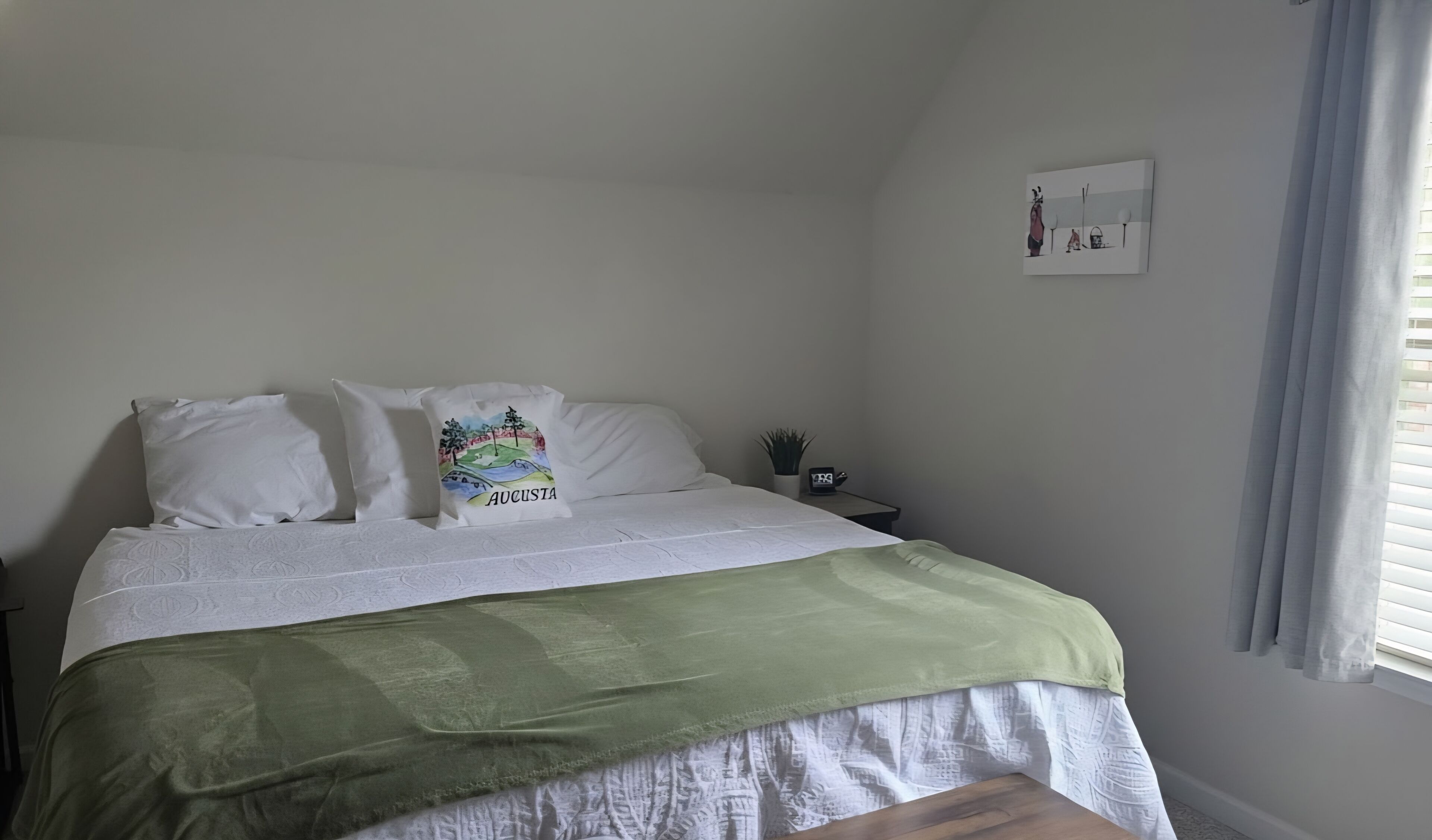 Ein Schlafzimmer mit einem Bett, Kissen, einer grünen Decke, einem Nachttisch mit Pflanze und einem an der Wand hängenden Bild.