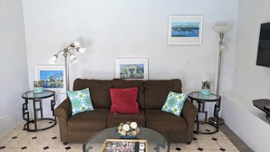 A living room with a brown sofa, two patterned pillows, a glass coffee table with decorative items, a framed painting on the wall, a floor lamp, and a small table with a lamp. - Sweet Dreams by the Pool - V (Smiths)
