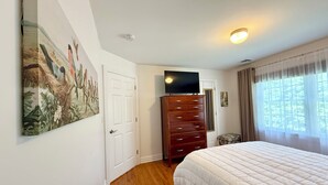 Une chambre avec un plancher en bois, un lit blanc, une commode, un téléviseur posé sur la commode et un tableau accroché au mur.