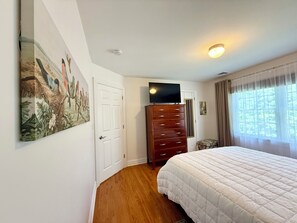 A bedroom with a wooden floor, a white bed, a dresser, a television on top of the dresser, and a painting on the wall. - NEW" Cozy pristine home in Union County NJ, Mins to NYC free coffee, Smart Tvs (Mountainside)