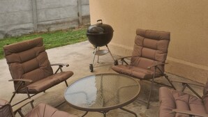 Une terrasse extérieure avec quatre chaises marron à coussins et une table ronde en verre, avec un barbecue noir en arrière-plan.