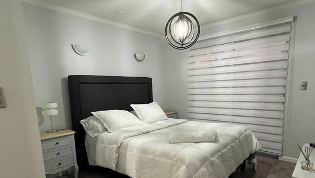 Une chambre à coucher comprenant un lit, des tables de chevet, une commode, un luminaire suspendu et une fenêtre avec des stores.