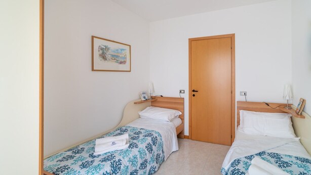 A small, clean room with two single beds, a wooden door, a small shelf with a lamp, and a framed picture on the wall. - Fabulous Apartment With Premium Amenities (San Michele al Tagliamento)