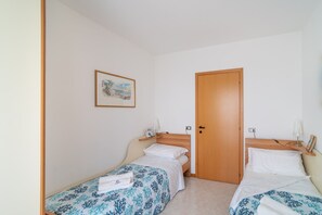 Una habitación pequeña y limpia con dos camas individuales, una puerta de madera, un estante con una lámpara y un cuadro en la pared.