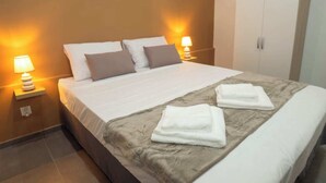 A neatly made bed with white and brown bedding, two bedside lamps, and a wardrobe in a hotel room. - Ixora (La Trinité)