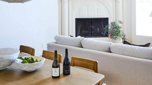 Un coin repas avec une table en bois, deux bouteilles de vin et un saladier de fruits. Une grande tapisserie est accrochée au-dessus de la cheminée, et un canapé est placé devant.
