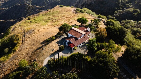 An aerial view of a house with a red-tiled roof, surrounded by a landscaped garden and trees, set in a hilly landscape.