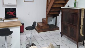 Een woonkamer met een houten trap, een keukenhoek met een witte koelkast en een televisie, en een bank met een plaid.
