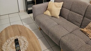 Een woonkamer met een grijze bank, een houten salontafel met een afstandsbediening, en een zwarte boekenkast tegen een witte muur.