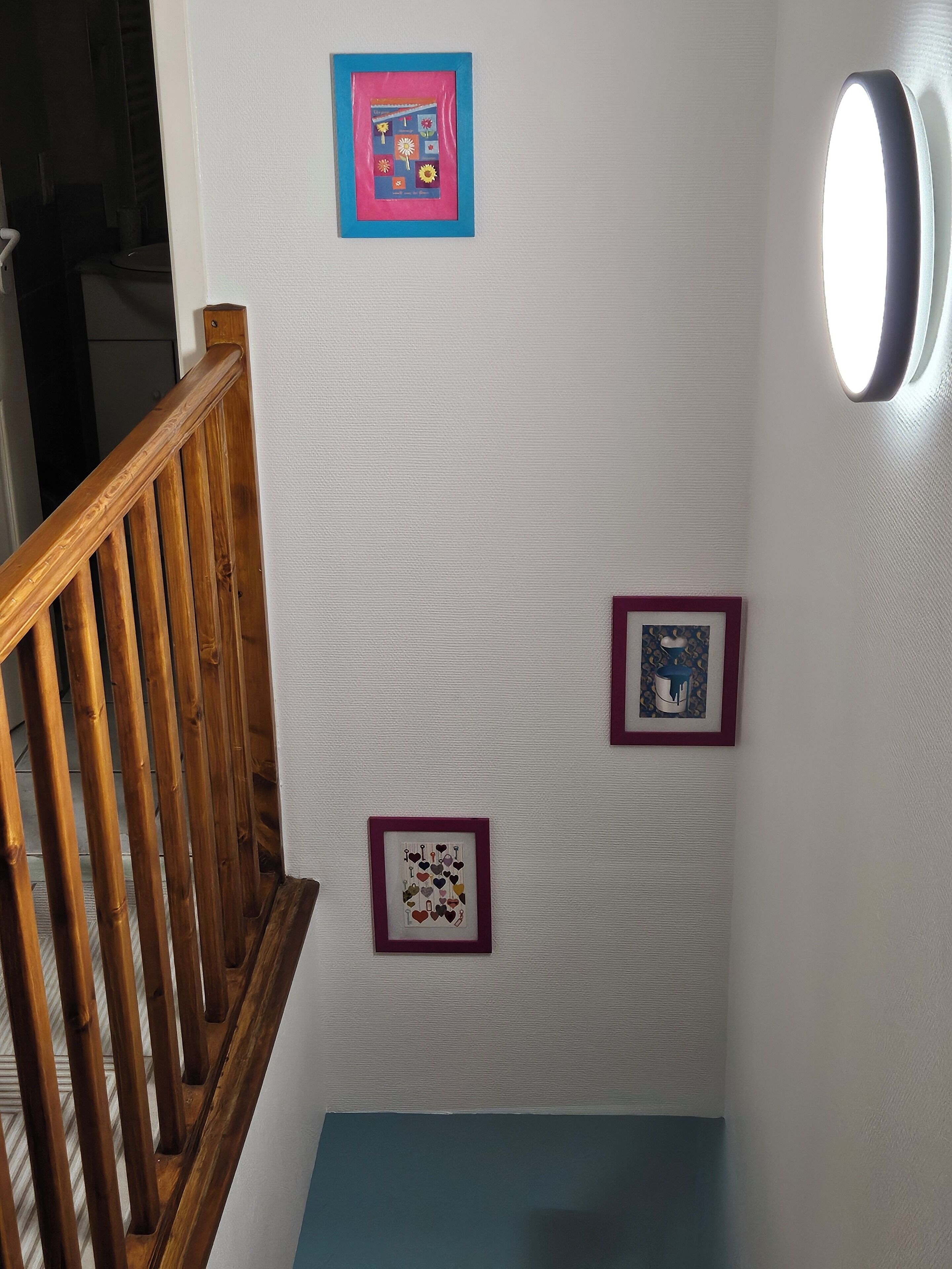 Una escalera con barandilla de madera, una pared con tres cuadros enmarcados y una lámpara fijada a la pared.