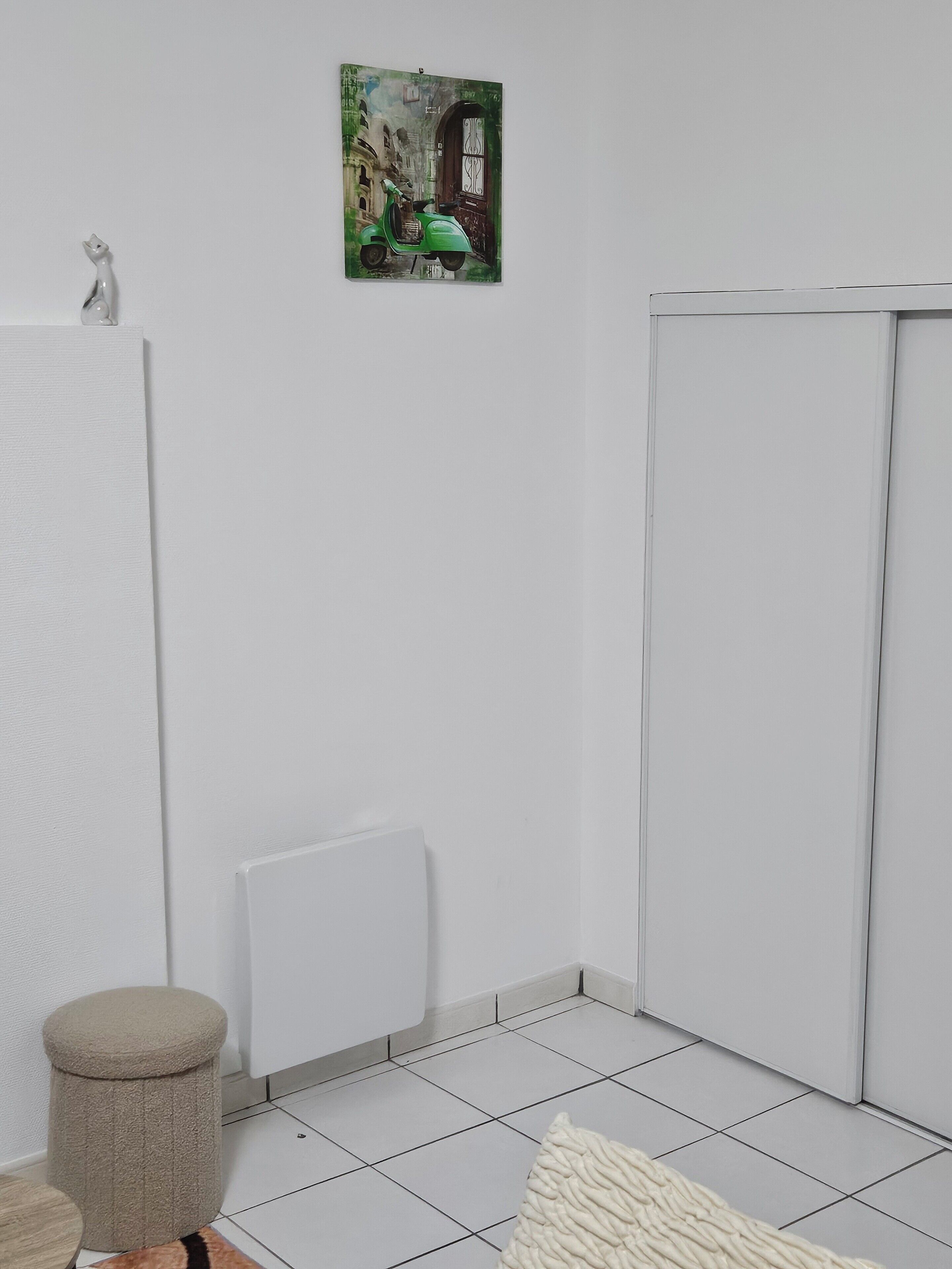 Un baño con un radiador blanco, un taburete color beige y un cuadro de una scooter verde en la pared.