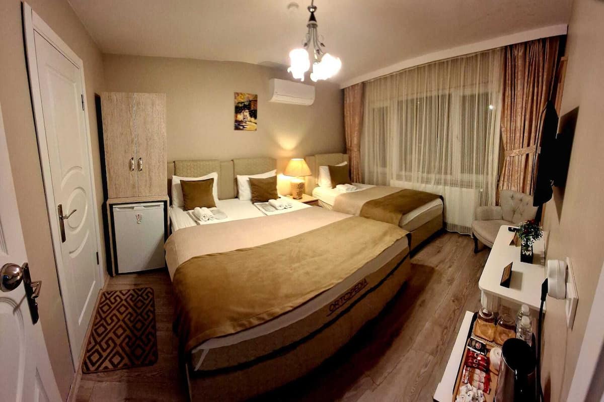 A hotel room with a large bed, bedside tables, a desk, a chair, a small fridge, and a window with curtains.