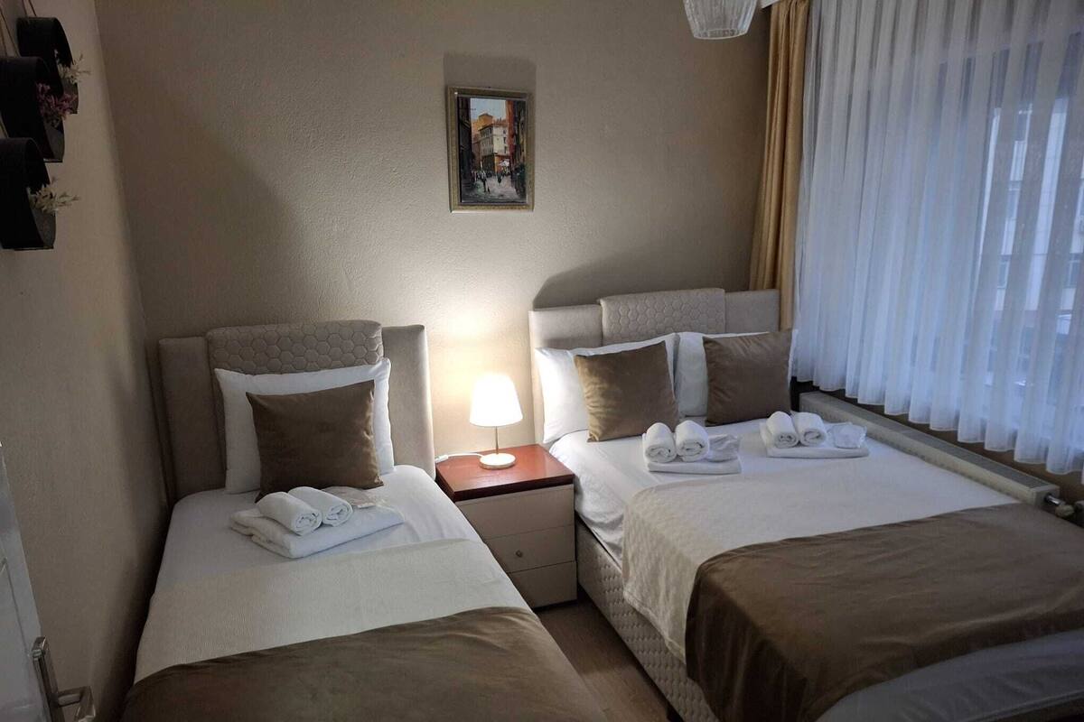 A hotel room with two beds, a nightstand with a lamp, a painting on the wall, and a window with curtains.