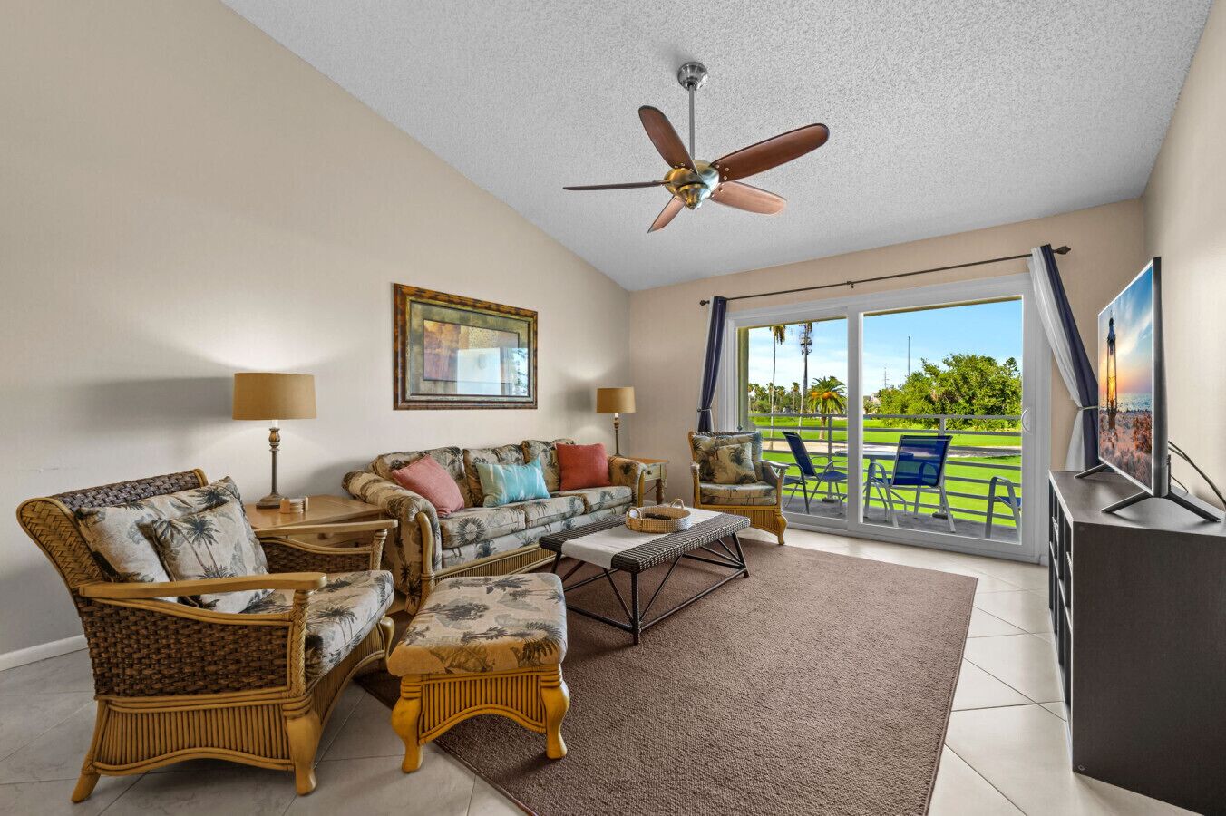 A spacious living room with a sofa, chairs, a coffee table, and a TV. There is a ceiling fan, a painting on the wall, and a view of a green landscape through the glass door.