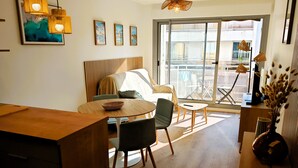A modern living room with a wooden dining table, a sofa, and a large window providing a view of the outside. - LA ROSE DES SABLES - Near Beach (Les Sables-d´Olonne)