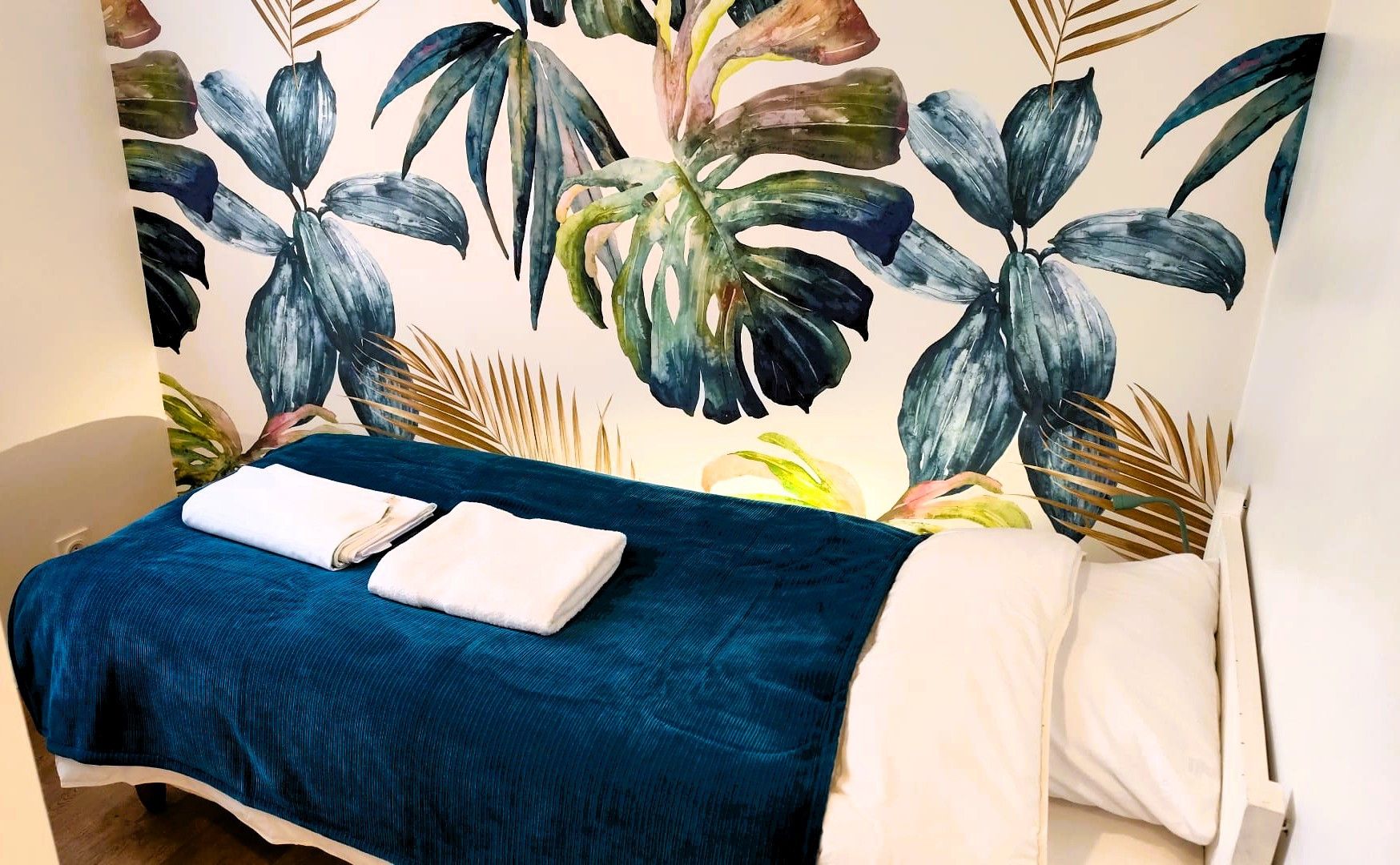 Quarto de hotel com uma cama, um cobreleito azul e uma parede com um mural de folhagem tropical.