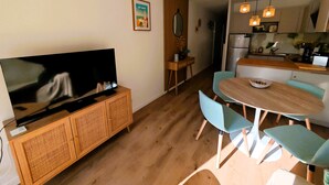 A modern living room with a flat-screen TV on a wooden cabinet, a dining area with a round table and blue chairs, and a kitchen with a refrigerator and hanging lights. - LA ROSE DES SABLES - Near Beach (Les Sables-d´Olonne)