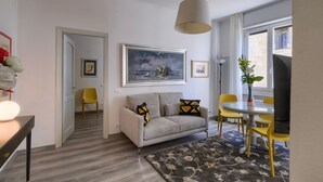 Een woonkamer met een bank, een eettafel met stoelen, een groot raam en een schilderij aan de muur.