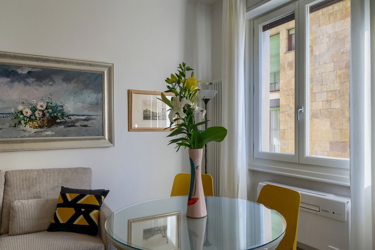 Een woonkamer met een glazen tafel, een lijstwerk met schilderij, een vaas met bloemen en uitzicht op een gebouw door het raam.