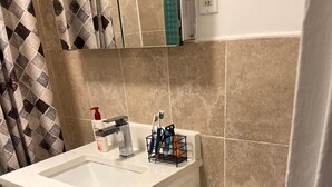 Baño con lavamanos blanco, inodoro y zona de ducha. Hay un portalapinceles en el lavamanos con cepillos de dientes.