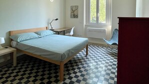 A bedroom with a bed, a desk, a chair, a window, and a radiator.