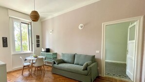 A living room with a herringbone wooden floor, a green sofa, a white table with chairs, a hanging light fixture, and a window with a view of trees.